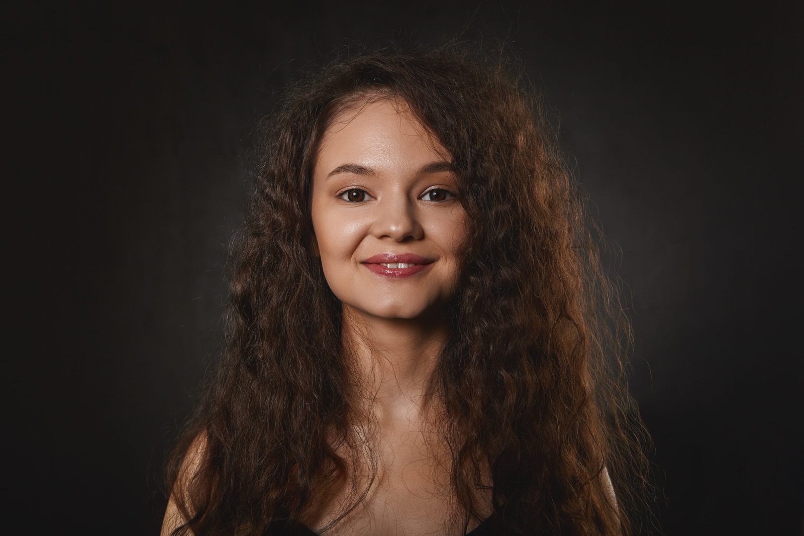 Charming cheerful young brunette lady with curly voluminous hairdo and perfect skin smiling happily, glad with good positive news, expressing joy. Pretty girl with dark loose hair posing in studio